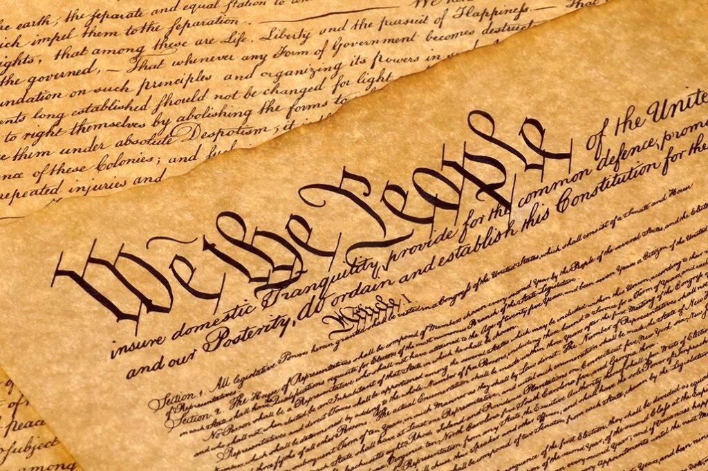 An image of an ancient parchment document displaying the text 'We The People' prominently at the top, with handwritten cursive and printed text, representing a foundational American political document.