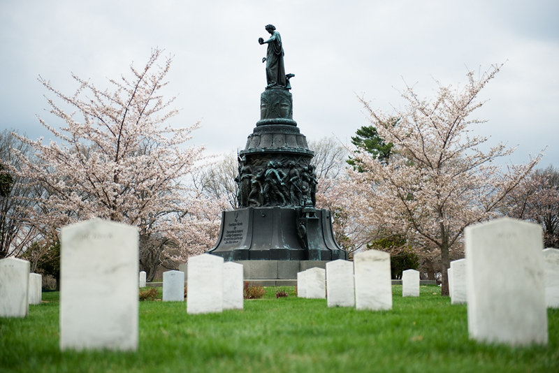 A statue on a pedestal surrounded by white gravestones and pink cherry blossom trees in Arlington Cemetery.