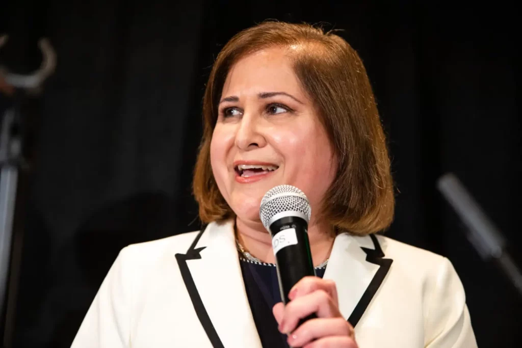 A woman speaking into a microphone, wearing a white jacket with black trim, appearing engaged and confident.