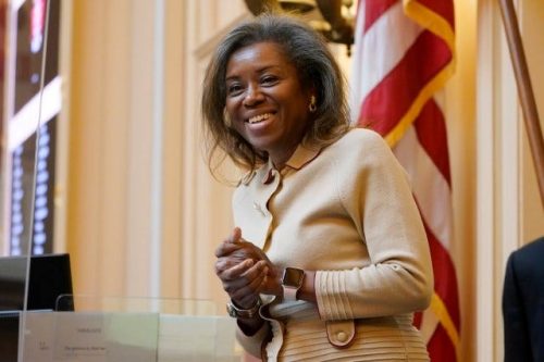 A smiling woman wearing a cream-colored suit stands in a legislative chamber, with an American flag in the background.