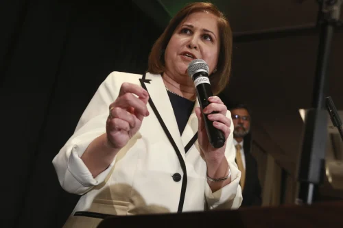 A woman in a white blazer speaks passionately into a microphone at a political event.