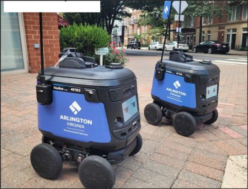Two small robots with the Arlington, Virginia logo parked on a brick sidewalk in an urban area.