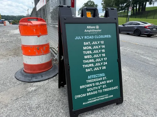 A roadside sign listing July road closures at Allianz Amphitheater, with dates and affected streets, alongside a construction cone.