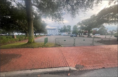 An empty parking lot with barricades set up, surrounded by trees and paving, with a building visible in the background.