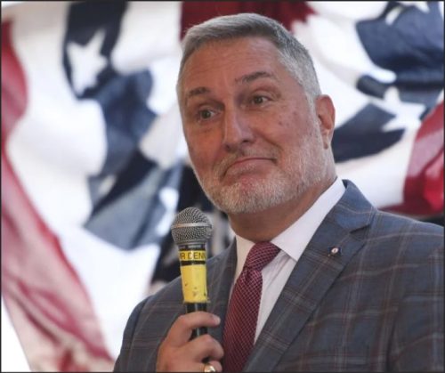 A man with a beard and short hair speaks into a microphone, wearing a suit and tie, with an American flag backdrop behind him.