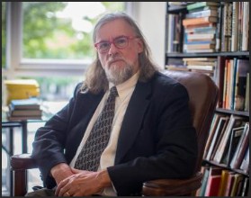 A man with long hair and glasses sits in a chair in front of a bookshelf filled with books. He is wearing a suit and tie, looking directly at the camera with a thoughtful expression.