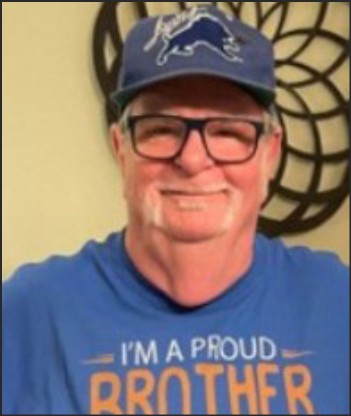 A smiling older man wearing a blue baseball cap with a lion logo, glasses, and a blue shirt that reads 'I'M A PROUD BROTHER.'