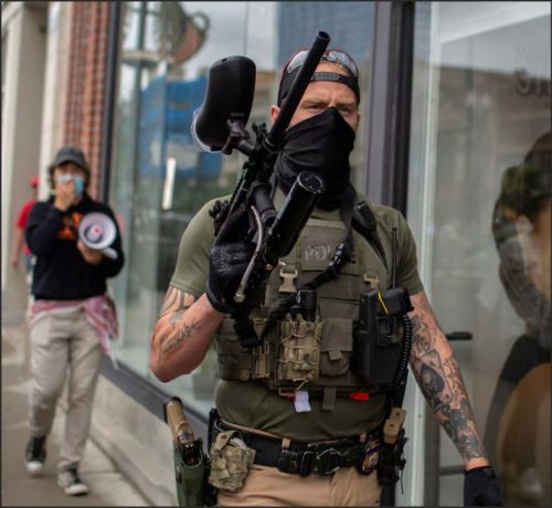 A person wearing tactical gear and a mask holds a paintball gun, walking past a storefront, while another individual behind them carries a megaphone.