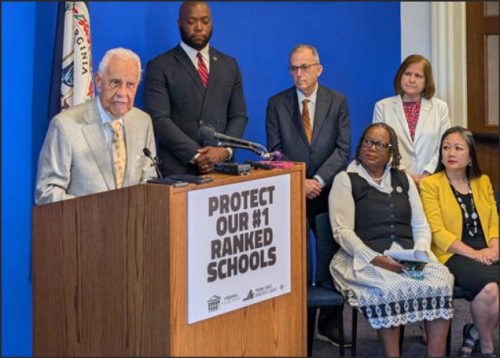 A group of politicians standing behind a podium with a sign that reads 'PROTECT OUR #1 RANKED SCHOOLS'. One elderly man in a suit speaks while others listen attentively.