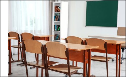 A classroom featuring wooden desks and chairs arranged in rows, with a green chalkboard and a bookshelf in the background.
