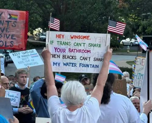 A protestor holds a sign reading 'HEY WINSOME, IF TRANS CAN'T SHARE YOUR BATHROOM THEN BLACKS CAN'T SHARE MY WATER FOUNTAIN' during a demonstration outside a school board meeting, surrounded by other demonstrators carrying various signs.