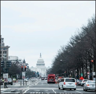 View of a city street leading to the U.S. Capitol building, lined with vehicles and trees under a cloudy sky.