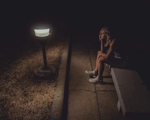 A young woman sits on a bench in a dimly lit area near a lamp, looking pensive and contemplative.