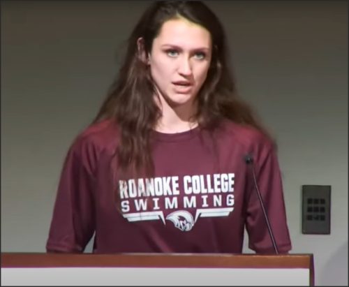 A young woman wearing a Roanoke College swimming shirt stands at a podium during a press conference.