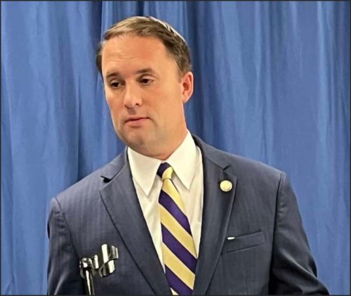 A man in a navy suit and striped tie standing in front of a blue backdrop, appearing to speak or address an audience.