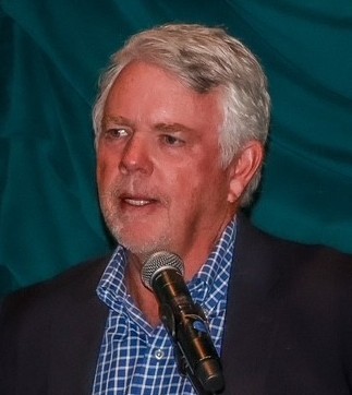 A man with grey hair and a beard is speaking into a microphone at a public event, wearing a blue checked shirt and a dark blazer, set against a teal backdrop.