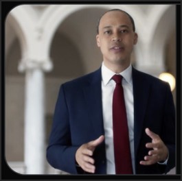 A man in a suit gestures while speaking in a formal setting with architectural features in the background.