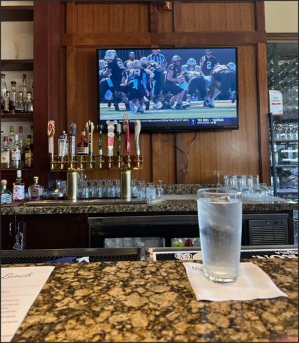 A bar counter with a glass of water in the foreground and a TV in the background displaying a college football game.