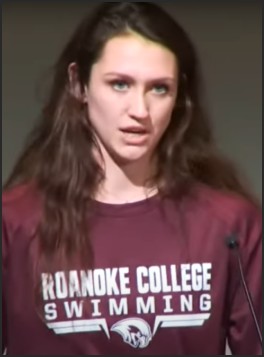 A young woman wearing a Roanoke College swimming shirt is speaking at a podium during a press conference, expressing her views.