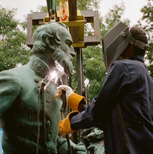 A person wearing protective gear appears to be working on the repair or removal of a bronze statue of a Confederate soldier, with equipment in the background.