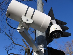 A close-up view of a traffic light equipped with a red light running camera, mounted on a pole against a clear blue sky.