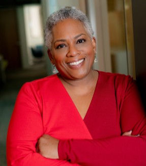 A smiling woman with short gray hair and a red dress is standing with her arms crossed, exuding confidence and warmth.