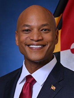Headshot of Maryland Governor Wes Moore smiling, wearing a suit and red tie, with the Maryland state flag in the background.