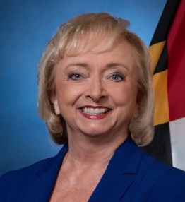 A woman with blonde hair and a blue blazer smiles in front of a blue background and a flag.