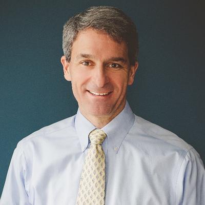 Portrait of Ken Cuccinelli, a man with short gray hair, wearing a light blue dress shirt and a patterned tie, smiling against a dark blue background.