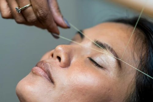 A close-up of a woman with closed eyes, having her eyebrows threaded by a person's hand using thin thread.