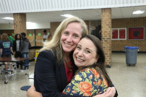 Two women embrace joyfully in a school cafeteria, with one wearing a blazer and the other adorned in a floral top.