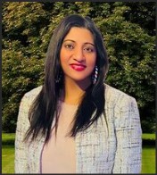 Portrait of a woman with long dark hair, wearing a light-colored top and a textured white blazer, smiling in front of a lush green background.