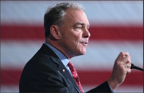 Senator Tim Kaine speaking at an event with an American flag in the background.