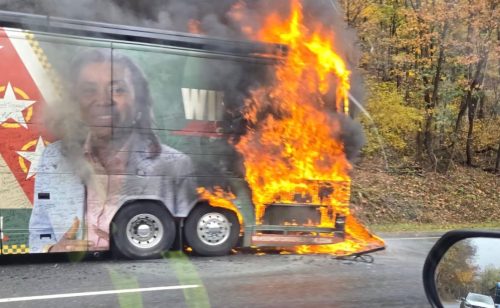 A campaign bus engulfed in flames on the side of the road, with a visible image of a candidate on its side.