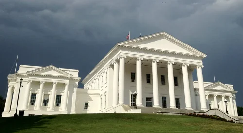 A view of the Virginia State Capitol building featuring its iconic white columns and clean architectural lines, set against a dramatic sky.
