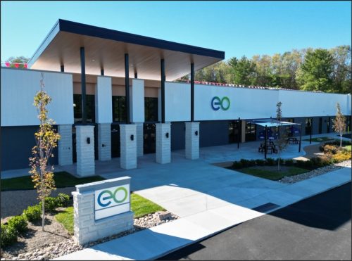 Modern building with a sleek design, featuring large windows, a well-lit entrance, and greenery in the surroundings. The structure prominently displays a logo on the front.