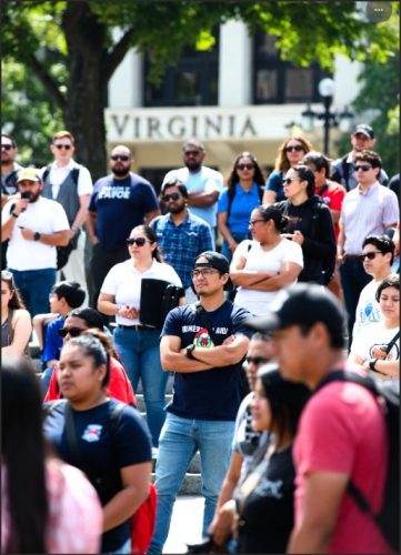 A diverse crowd of people gathered outdoors in Virginia, with some participants wearing casual clothing and expressions of engagement, amidst trees and a building labeled 'Virginia' in the background.