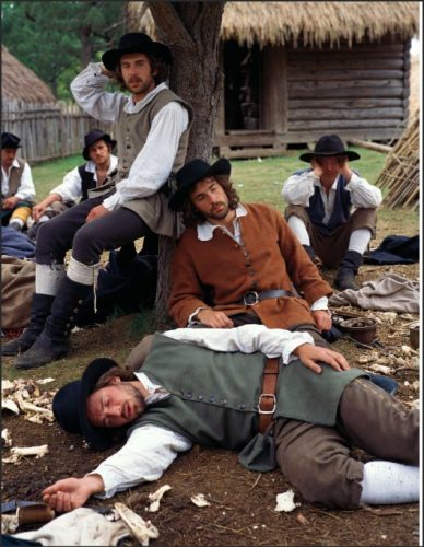 Group of historical reenactors dressed in 17th-century attire, sitting and resting under a tree near a rustic wooden settlement.