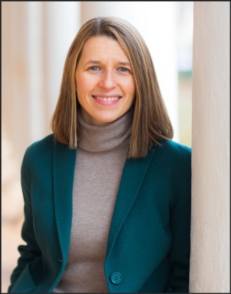 A professional headshot of a woman with shoulder-length brown hair, wearing a green blazer over a beige turtleneck, smiling while leaning against a column.