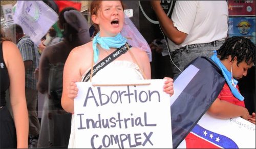 A protester passionately holding a sign that reads 'Abortion Industrial Complex,' wearing a blue bandana and surrounded by other demonstrators.