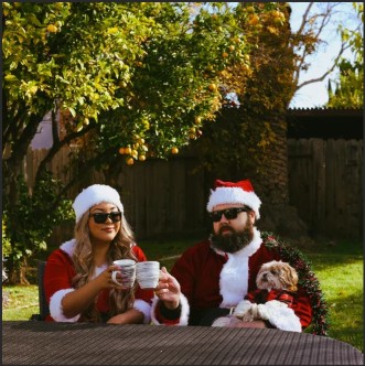 A man and a woman in Santa costumes sit at a table outdoors, holding mugs and wearing sunglasses. A small dog is sitting in the man's lap, and there are orange trees in the background.