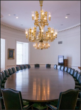 A large, oval wooden conference table surrounded by black leather chairs, illuminated by an ornate chandelier in a well-lit room.