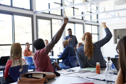 A classroom setting with students actively participating; some are raising their hands while a teacher engages with them.