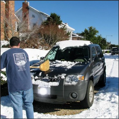 A person shoveling snow off the hood of a gray SUV parked in a snowy residential area.
