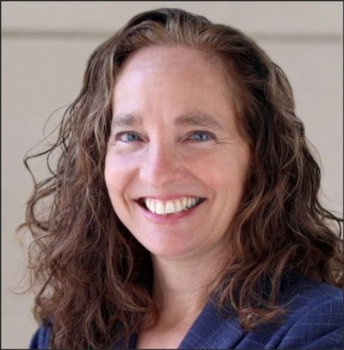 A smiling woman with curly brown hair, wearing a dark blazer, poses for a portrait against a neutral background.