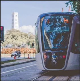 A modern tram approaching a stop on tracks, with a tower in the background and trees lining the street.