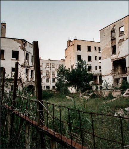 An abandoned building complex with crumbling walls and broken windows, surrounded by overgrown grass and a rusty fence.