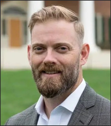 A man with short, light brown hair and a full beard smiles at the camera. He is wearing a gray suit jacket over a white shirt, with a blurred green lawn and building in the background.