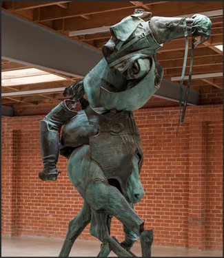A bronze sculpture depicting a horse rearing up with a knight in armor, set against a brick wall in an art gallery.