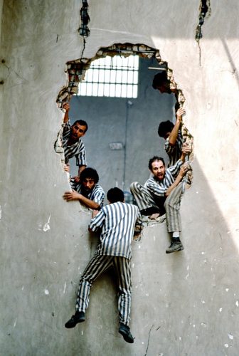 Group of prisoners in striped uniforms climbing through a large hole in a wall.